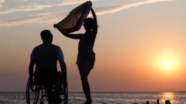 Uzun bacaklı kadın iskelede duruyor ve kötürüm bir adamın önünde bez sallıyor. Arka planda tekerlekli sandalyeye mahkum.