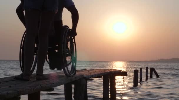Gondoskodás a kerekesszékben ülő személy, aki élvezi a gyönyörű naplemente a víz felett — Stock videók