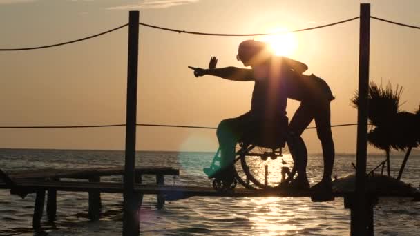 Chanceux handicapés en fauteuil roulant avec femme regardant à l'horizon sur fond de beau coucher de soleil au voyage d'été — Video