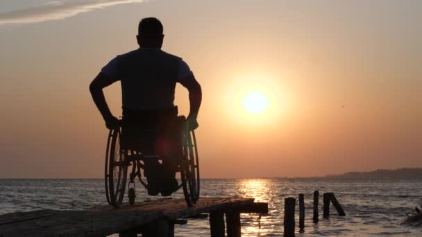 Handicapé mâle en fauteuil roulant reposant sur le fond de ciel orange et l'eau au coucher du soleil — Video