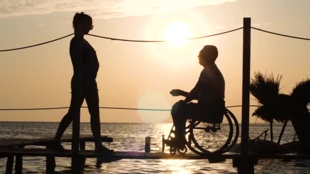 Vergadering van slank meisje met een handicap jongen op rolstoel bij heldere zonsondergang staan op pier in de buurt van water — Stockvideo