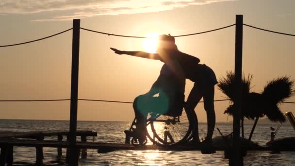 Tevreden gehandicapt op rolstoel praten met vrouwen en kijken in afstand op de achtergrond van zonsondergang op zee — Stockvideo