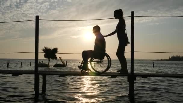 Mooi vrouw met man ongeldig op rolstoel gaan op steiger tegen de hemel in zonsondergang — Stockvideo