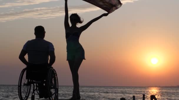 Shapely girl is standing on jetty and waving cloth in front of man disabled into wheel chair on background of sunset at summer weekend — Stock Video