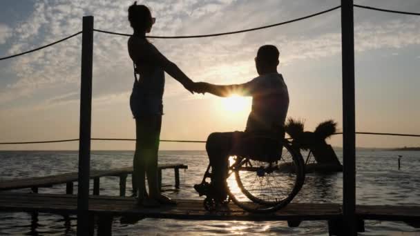 Silhouette von Verliebten im Rollstuhl am Kai vor dem Hintergrund von Wasser und Himmel bei Sonnenuntergang — Stockvideo