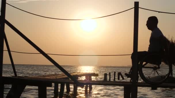 Silhouette de l'homme avec des pieds douloureux sur fauteuil roulant sur jetée profitant du coucher de soleil sur la mer au voyage d'été — Video