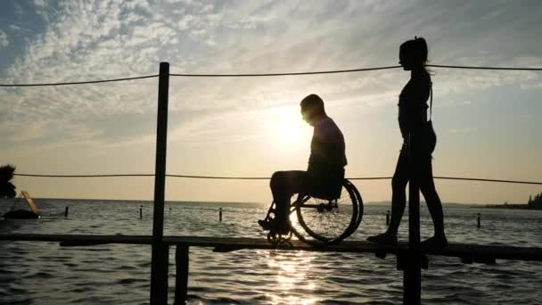 Esbelta chica con hombre discapacitado en silla de ruedas caminando en el muelle en el mar contra el cielo en la puesta del sol — Vídeos de Stock