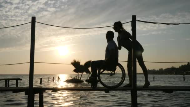 Gehandicapte die een volledig leven leidt jonge vrouwen met mannetjes kreupel op rolstoel naar pier aan de waterkant op de achtergrond hemel in oranje zonnestralen — Stockvideo
