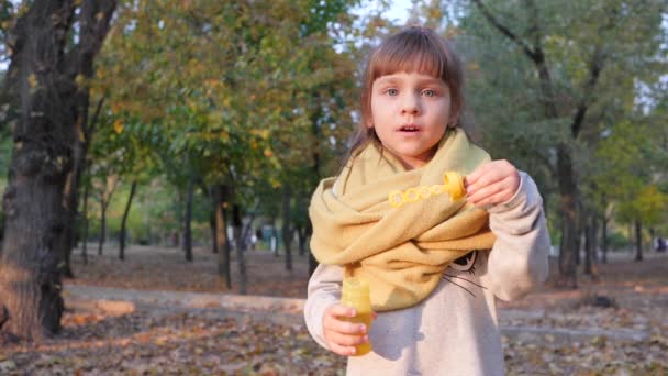 Щаслива маленька дівчинка дме повітряні бульбашки і посміхається в парку на фоні дерев — стокове відео