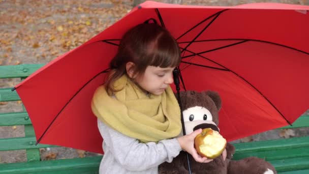 Маленькая девочка и друг-игрушка сидят на скамейке в парке под зонтиком и едят красное яблоко — стоковое видео
