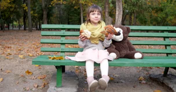 Дитина тримає два шматки булочки з маком, що сидить біля плюшевого ведмедя на лавці на дитячому майданчику в парку на відкритому повітрі — стокове відео