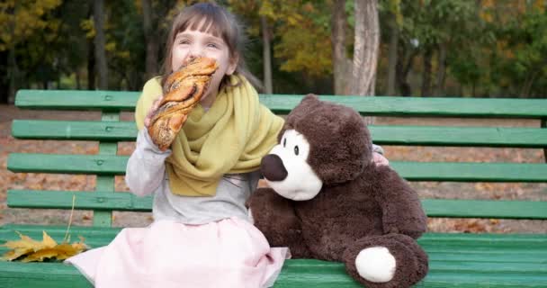 Niña come pan trenzado con semillas de amapola en el banco cerca de oso de peluche en el parque de otoño — Vídeos de Stock