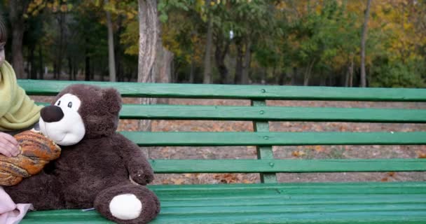 Kid eats sweet bun with poppy on bench with teddy bear toy at playground in autumn park outdoors — Stock Video