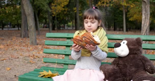 Малыш с двумя кусочками булочки с маком в руках сидит рядом с плюшевым мишкой на скамейке на детской площадке в осеннем парке — стоковое видео