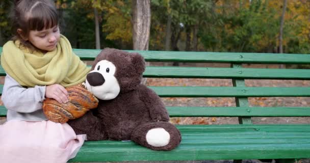 Маленька дівчинка їсть плетену булочку з маком на лавці з плюшевим ведмедем на дитячому майданчику в осінньому парку — стокове відео