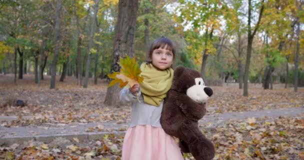 Petite fille marche avec ours en peluche et feuilles jaunes dans les mains au parc d'automne, jouer avec jouet à l'extérieur — Video