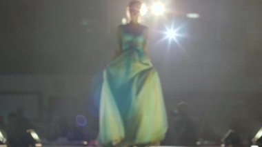 Moda gösterisinde şık elbiseler, genç mankenler podyumda insanların önünde poz veriyorlar.