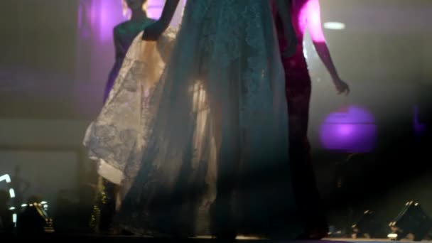 Модели в шикарных вечерних платьях крупным планом позируют на подиуме на фоне дыма и света — стоковое видео