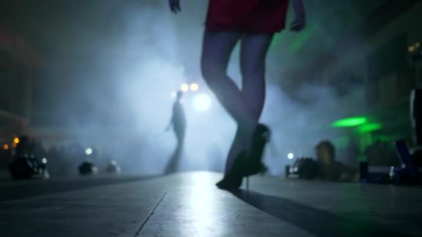 ヒールの高い靴のデザイナー服や足の長いモデルのプレゼンテーションは煙の中でサーチライトに照らされた表彰台に沿って行く — ストック動画