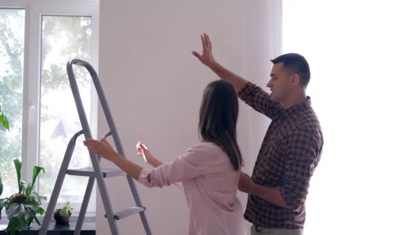 Mooi paar doen renovatie en met behulp van trapladder voor hangen foto op witte muur in platte — Stockvideo