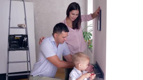 Papá enseña a hijo a atornillar estante a pared con destornillador y madre cerca de colgar imagen durante la renovación apartamento — Vídeo de stock