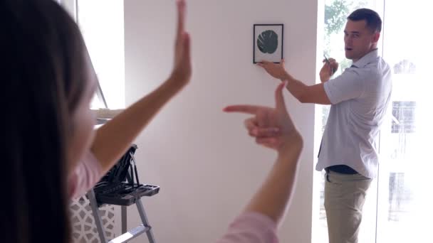 Decorate interior, attractive guy hangs picture while woman makes frame with finger and decide on decoration at new flat — Stock Footage Decorate interior, attractive guy hangs picture while woman makes frame with finger and decide on decoration at new flat Royalty Free Stock Footage