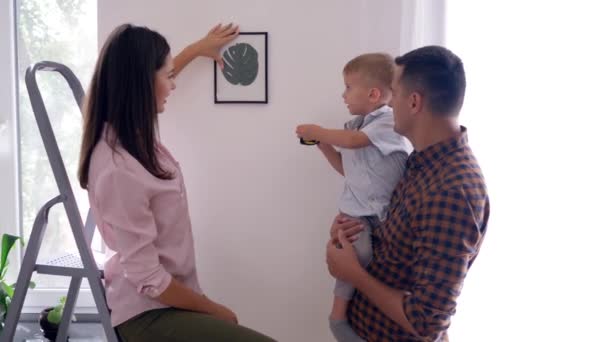 Decorare interni, bel bambino con roulette misurazione aiuta i genitori a fare le riparazioni in appartamento e appendere immagine — Video Stock
