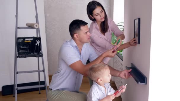 Familia amigable con niño pequeño haciendo redecorar y atornillar estante y foto a la pared — Vídeos de Stock