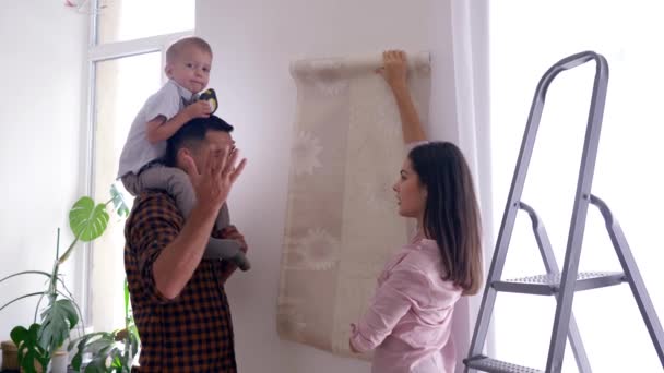 Счастливая семья делает ремонт в новом доме, веселая мама с сынишкой на плечах выбирают обои в квартире — стоковое видео