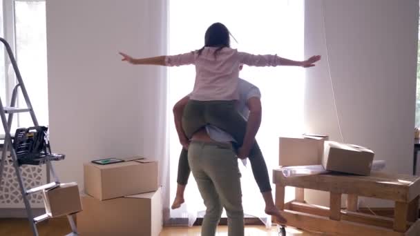 Feliz mudanza a nuevo apartamento, alegre pareja se divierte y el hombre está llevando a la mujer en la espalda durante el calentamiento de la casa — Vídeos de Stock