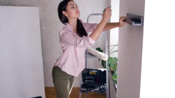 Счастливая молодая женщина украшает интерьер и вешает полку на стену во время ремонта новой квартиры — стоковое видео