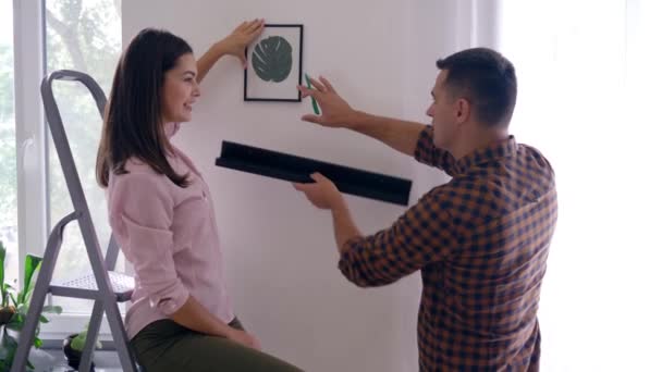 Home renovation, happy newlyweds hang painting and shelf on wall and girl kisses male in apartment — Stock Video