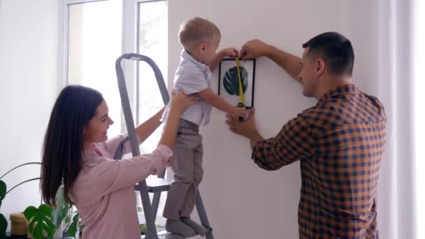 Reparación del hogar, lindo niño con cinta métrica ayuda a los padres a hacer reparaciones y colgar la imagen en el apartamento — Vídeo de stock