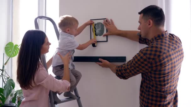 Niño bonito con cinta métrica ayuda a madre y padre colgar estante con imagen en la pared después de la reparación en plano — Vídeos de Stock