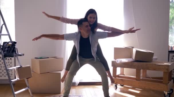 Mudança para nova habitação, par alegria está rindo e cara está carregando menina de volta entre as caixas durante a mudança para o apartamento — Vídeo de Stock