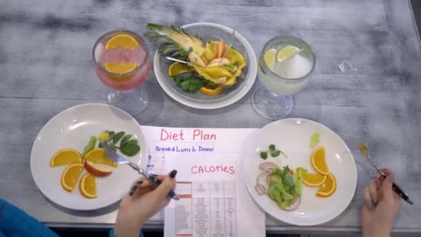 Фитнес-диета, девочки едят овощи и фрукты из тарелок, сидя за столом с калориями стол и план диеты — стоковое видео