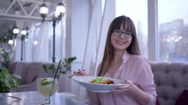 Хорошее питание в кафе, портрет молодой женщины в очках с вилкой и тарелкой в руке, поедающей греческий салат и смотрящей в камеру — стоковое видео