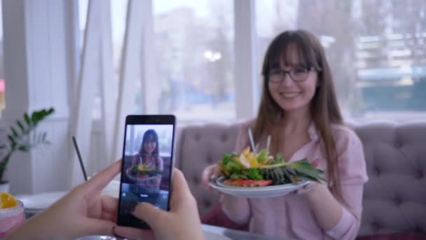 Sağlıklı beslenme ve blog yazma, gözlüklü neşeli kız tabağında taze meyvelerle kafedeki sosyal ağlar için cep telefonuyla fotoğraf çeken kız arkadaşına poz veriyor. — Stok video