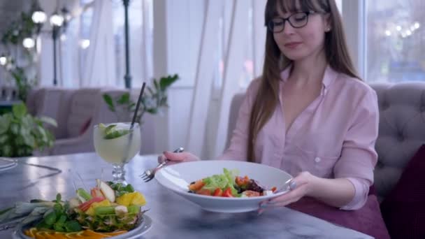Здоровый образ жизни, девушка с длинными волосами в очках с вилкой и тарелкой в руке ест греческий салат и смотрит в камеру — стоковое видео