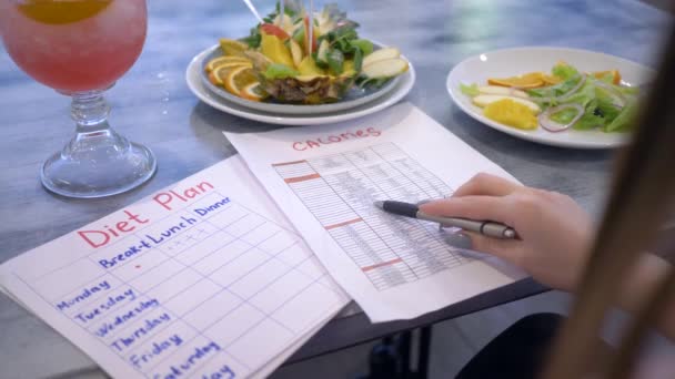 Ernährungswissenschaftlerin weiblich auf der Suche nach Kalorien in Liste und halten in der Hand Diätplan sitzt am Tisch mit Punsch und Obst in Teller — Stockvideo