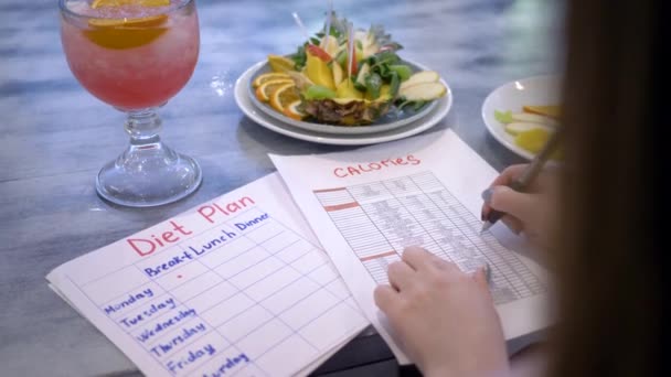 Nutritionist woman writes diet plan with food calorie sheet on table with punch and fruits in plate at cafe — Stock Video