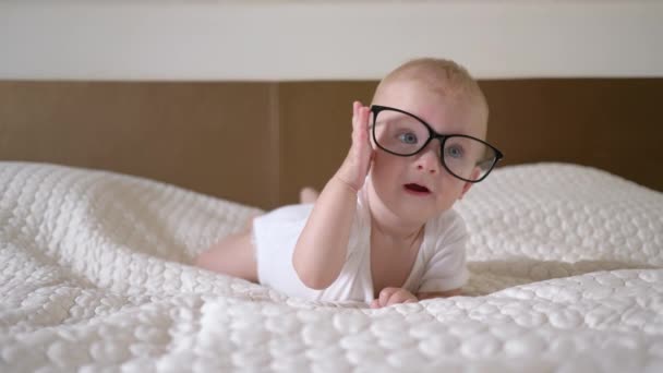 Дитинство, портрет милого маленького хлопчика з великими блакитними очима в окулярах лежить на ліжку крупним планом — стокове відео