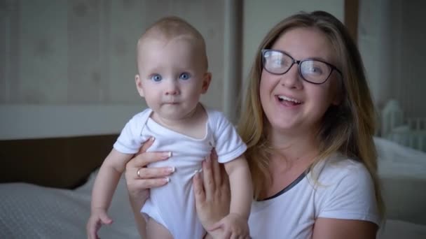 Família feliz, mãe alegre em óculos segura bebê a mãos e olha na câmera de perto no quarto — Vídeo de Stock