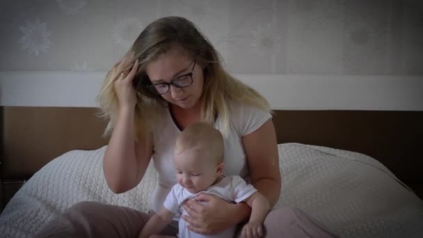 Уставшая расстроенная женщина с головной болью держит ребенка на руках, сидя на кровати дома — стоковое видео