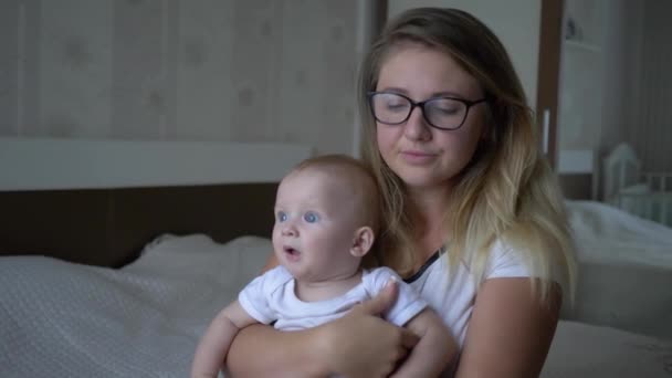 Несчастная уставшая женщина держит мальчика на руках и смотрит в камеру, сидя на кровати дома — стоковое видео
