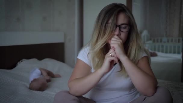 Worried woman in eyeglasses suffers from headachesits on background of baby on bed at home — Stock Video