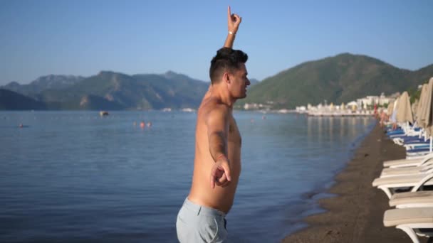 Ευτυχισμένος άνθρωπος χαζολογάει στην παραλία, χορεύει και πηδάει κοντά στη θάλασσα σε ένα φόντο βουνών και γαλάζιου ουρανού — Αρχείο Βίντεο