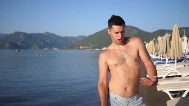 Homem feliz na praia, movimento engraçado perto do mar contra o fundo das montanhas e céu azul — Vídeo de Stock