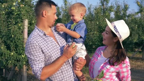 Criança alegre bebe o suco de maçã com pais no fundo de árvores, família feliz de agricultores no jardim — Vídeo de Stock