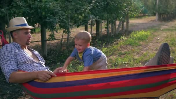 Счастливые моменты, ребенок пожимает отцу руку в гамаке возле фруктовой корзины в яблоневом саду — стоковое видео